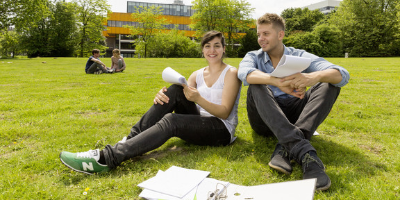 Zwei Studierende sitzen auf einer Wiese, im Hintergrund die Mensa
