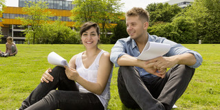 Zwei Studierende sitzen auf einer Wiese, im Hintergrund die Mensa