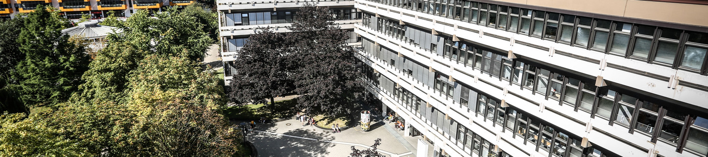 Emil-Figge-Straße 50: Ein langes Gebäude mit Fenstern, vor denen teilweise Rollos heruntergezogen sind, von oben fotografiert. Davor ein Platz mit Bäumen. Hinten links im Hintergund das gelb-weiße Mensa-Gebäude.